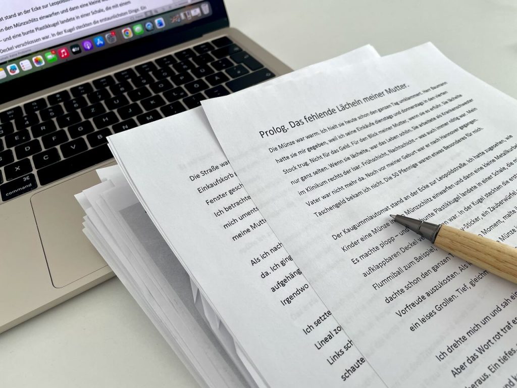 Gedrucktes Manuskript mit Prolog-Seite auf einem Schreibtisch neben einem Laptop und einem Stift.
